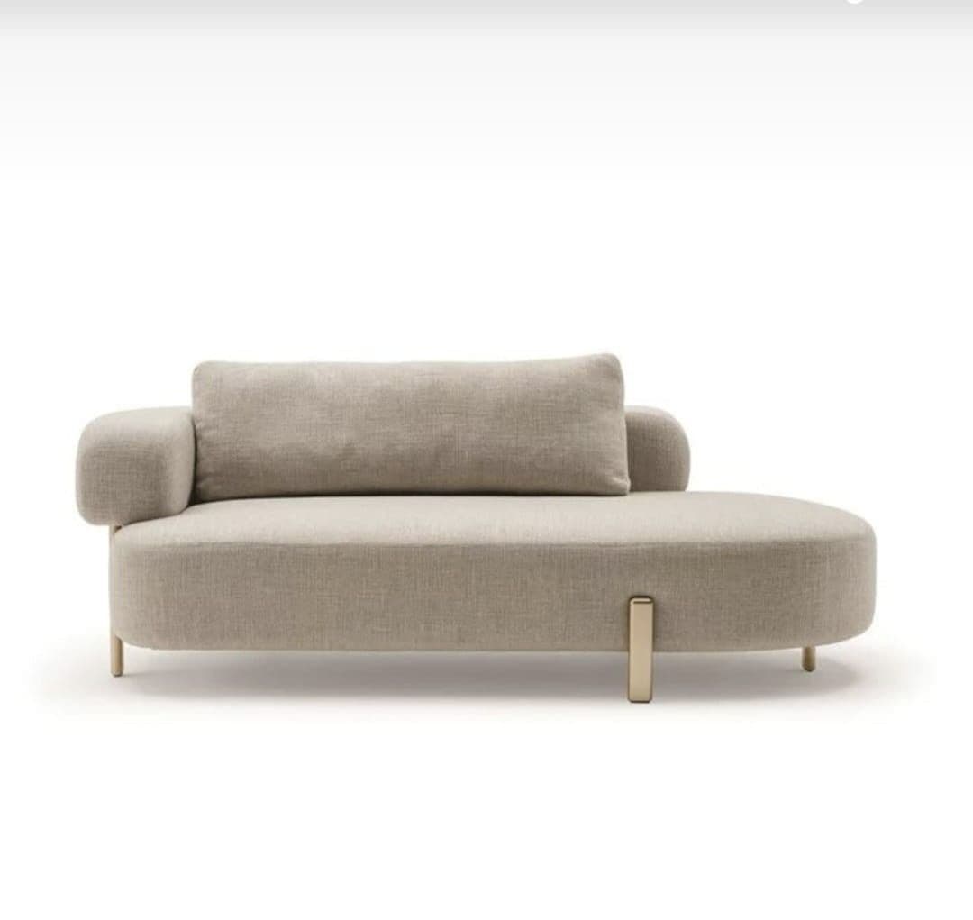 A contemporary chaise lounge with a rounded, upholstered form. It features a low profile, a single armrest on the left side that curves outward, and a long, rectangular back cushion. The piece is elevated by thin, angled legs, with a distinctive gold-toned metal accent piece visible on the front right side, extending upwards from the frame. — 3D model
