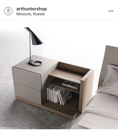 A modern, asymmetric nightstand featuring a two-drawer cabinet section in light gray and an integrated, lower open shelving unit in light wood. A small black conical table lamp rests on the top surface of the drawer unit, which is part of a wider base. — 3D model