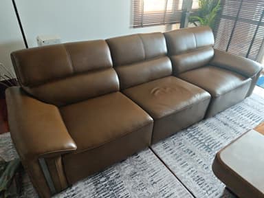 A contemporary three-seater sofa upholstered in rich brown leather, featuring distinct, segmented cushions suggesting individual reclining functionality. The wide armrests are gently curved and include subtle dark metallic accents on their outer sides, contributing to its modern aesthetic. The backrests are designed with an integrated headrest-like shape for enhanced comfort. — 3D model