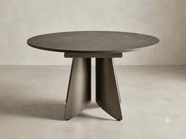 A modern round dining table featuring a dark, textured tabletop, which appears to be made from a sintered stone or ceramic material, supported by a distinctive, sculptural pedestal base crafted from dark bronzed metal. The tabletop shows a visible seam, suggesting it may be extendable. The base consists of four robust, trapezoidal legs that converge towards the center, providing stability and a contemporary aesthetic. — 3D model