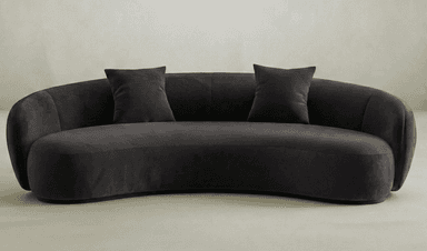 A contemporary curved sofa with a rounded silhouette. It features a low-profile design with integrated armrests that flow seamlessly into the backrest. The upholstery appears to be a soft fabric, possibly velvet or microsuede. Two matching throw pillows are placed on the sofa. The base is a dark, flat plinth. — 3D model