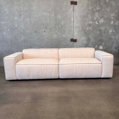 A modern, two-seater sofa with a low profile and blocky arms. The sofa is upholstered in a light-colored, textured fabric and features contrasting dark piping along the edges of the cushions and frame. The base appears to be a dark, solid plinth. It is positioned against a rough, concrete wall with visible imperfections and mounted fixtures. — 3D model