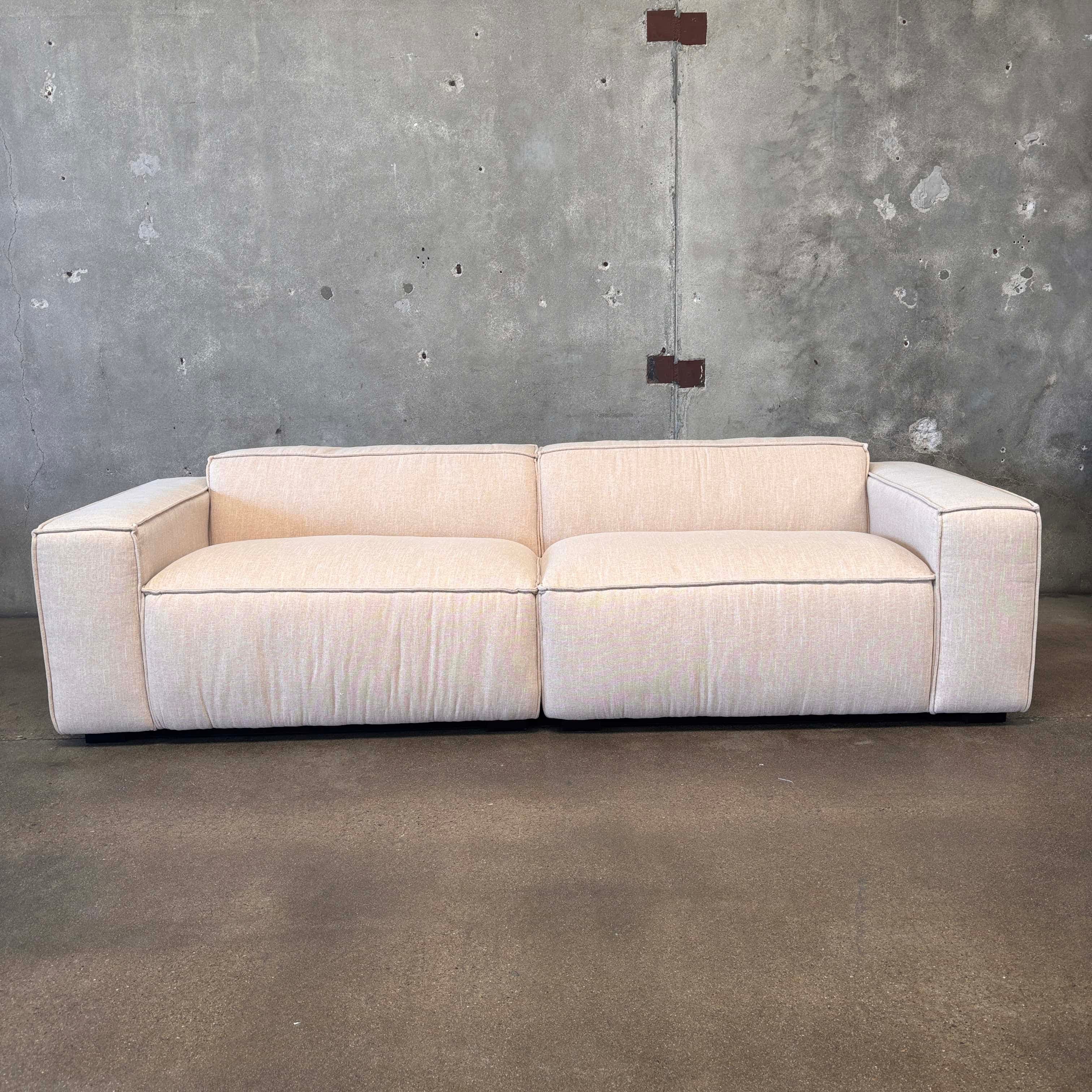 A modern, two-seater sofa with a low profile and blocky arms. The sofa is upholstered in a light-colored, textured fabric and features contrasting dark piping along the edges of the cushions and frame. The base appears to be a dark, solid plinth. It is positioned against a rough, concrete wall with visible imperfections and mounted fixtures. — 3D model
