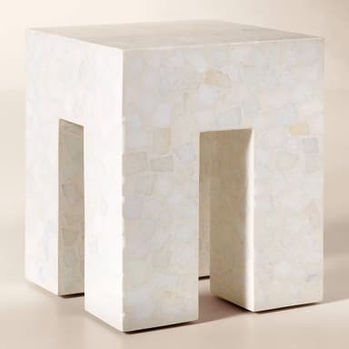 A monolithic cube-shaped side table crafted from light-toned terrazzo. The table features a solid top surface and two thick, rectangular legs formed by deep, symmetrical U-shaped cutouts on opposing sides, giving it an open yet sturdy appearance. The terrazzo material is composed of a light cream or off-white matrix embedded with irregular, lighter-colored aggregate chips, creating a speckled texture across all visible surfaces. — 3D model