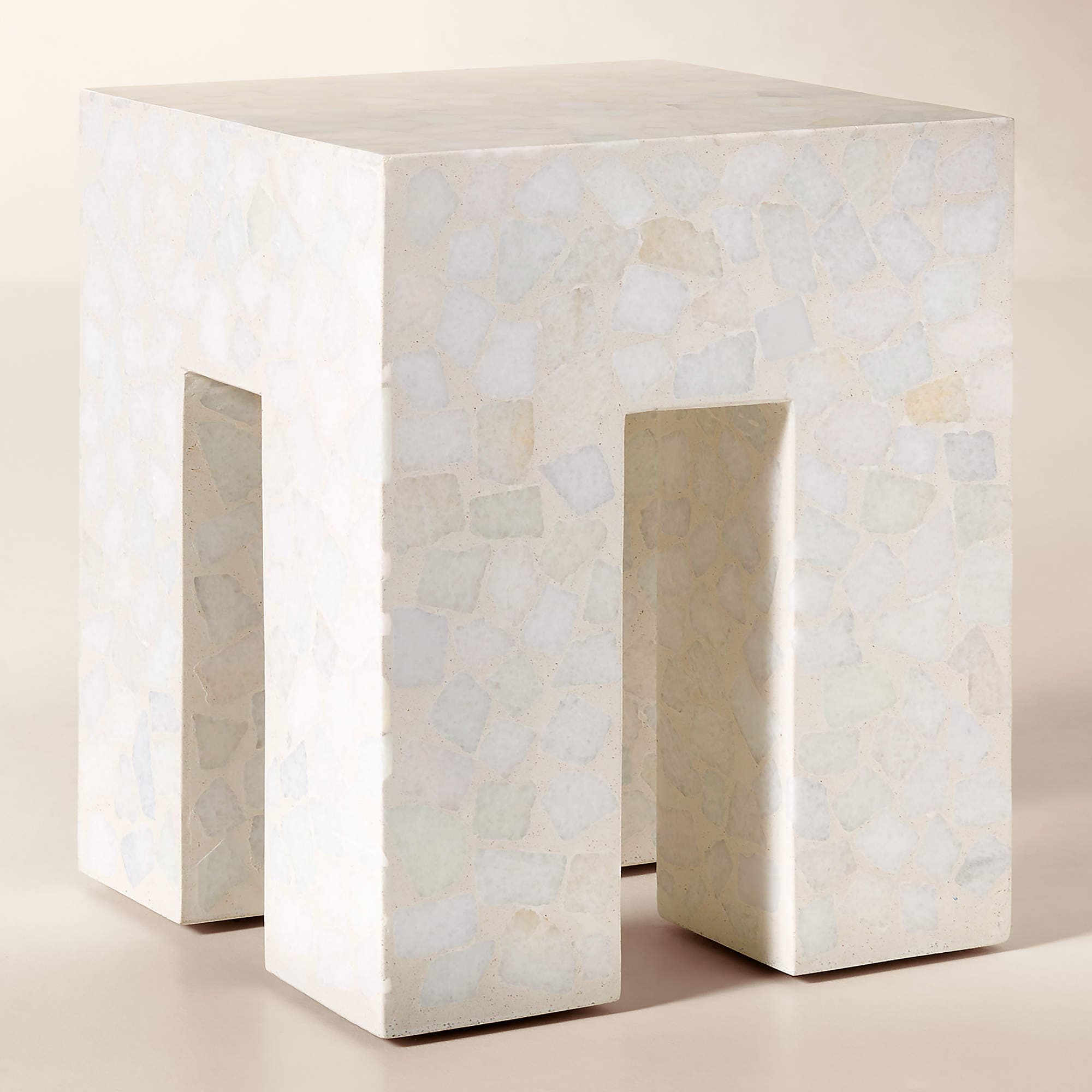 A monolithic cube-shaped side table crafted from light-toned terrazzo. The table features a solid top surface and two thick, rectangular legs formed by deep, symmetrical U-shaped cutouts on opposing sides, giving it an open yet sturdy appearance. The terrazzo material is composed of a light cream or off-white matrix embedded with irregular, lighter-colored aggregate chips, creating a speckled texture across all visible surfaces. — 3D model
