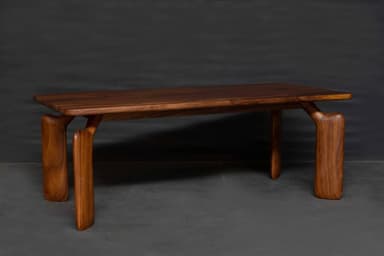 A low, rectangular bench crafted from dark-toned solid wood, likely walnut. It features a thick, smoothly finished top with softly rounded edges. The most striking design element is its four sculptural, curvilinear legs that appear to flow seamlessly from the underside of the bench top, creating an organic and monolithic form. The legs are wide and substantial, tapering slightly before flaring at the base, providing stable support. The overall aesthetic is minimalist yet impactful, highlighting the natural beauty and grain of the wood. — 3D model