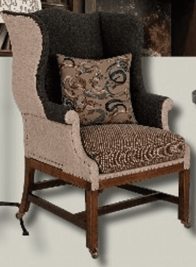 A wingback chair featuring a high back with distinctive 'wings', upholstered in a combination of two fabric types. The primary upholstery is a textured neutral fabric on the sides and lower back, with a darker, possibly charcoal-colored fabric on the upper back and arms. Decorative brass nailhead trim outlines the transition between fabrics and along the seat base. It includes a seat cushion and a decorative throw pillow. The chair is supported by a wooden frame with four legs, the front two of which are fitted with brass casters. The legs and structural supports are made of dark stained wood. — 3D model