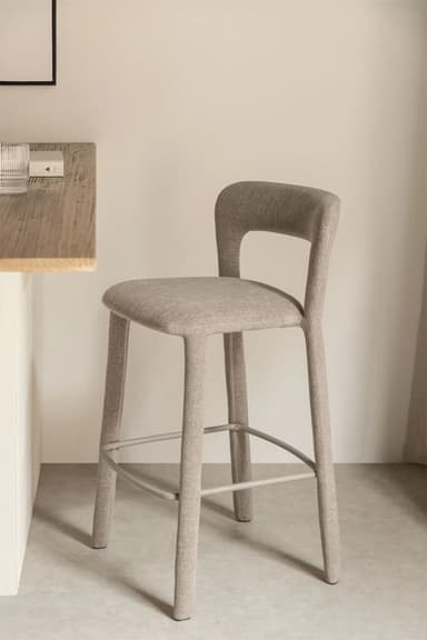 A modern bar stool with a slightly curved backrest and a seat upholstered in a textured beige fabric. The stool has four tapered legs that extend upwards to form the seat and backrest structure. Metal footrests are integrated between the legs. — 3D model
