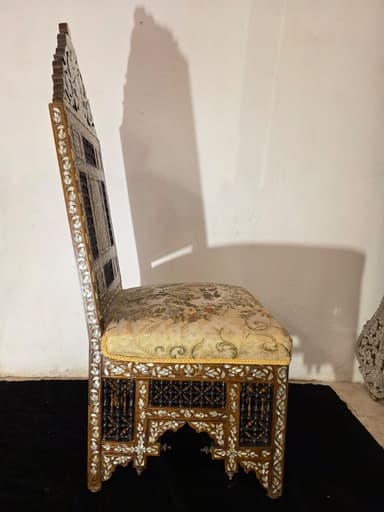 A highly ornate accent chair featuring intricate marquetry and inlay work. The chair has a tall, elaborately carved back with floral motifs and geometric patterns. The seat is upholstered in a light-colored fabric with a floral pattern. The base of the chair, including the legs and the area beneath the seat, is constructed from dark wood and heavily decorated with mother-of-pearl inlay, creating floral and geometric designs. The legs have an arched structure. The overall construction suggests a piece of antique or artisan craftsmanship. — 3D model