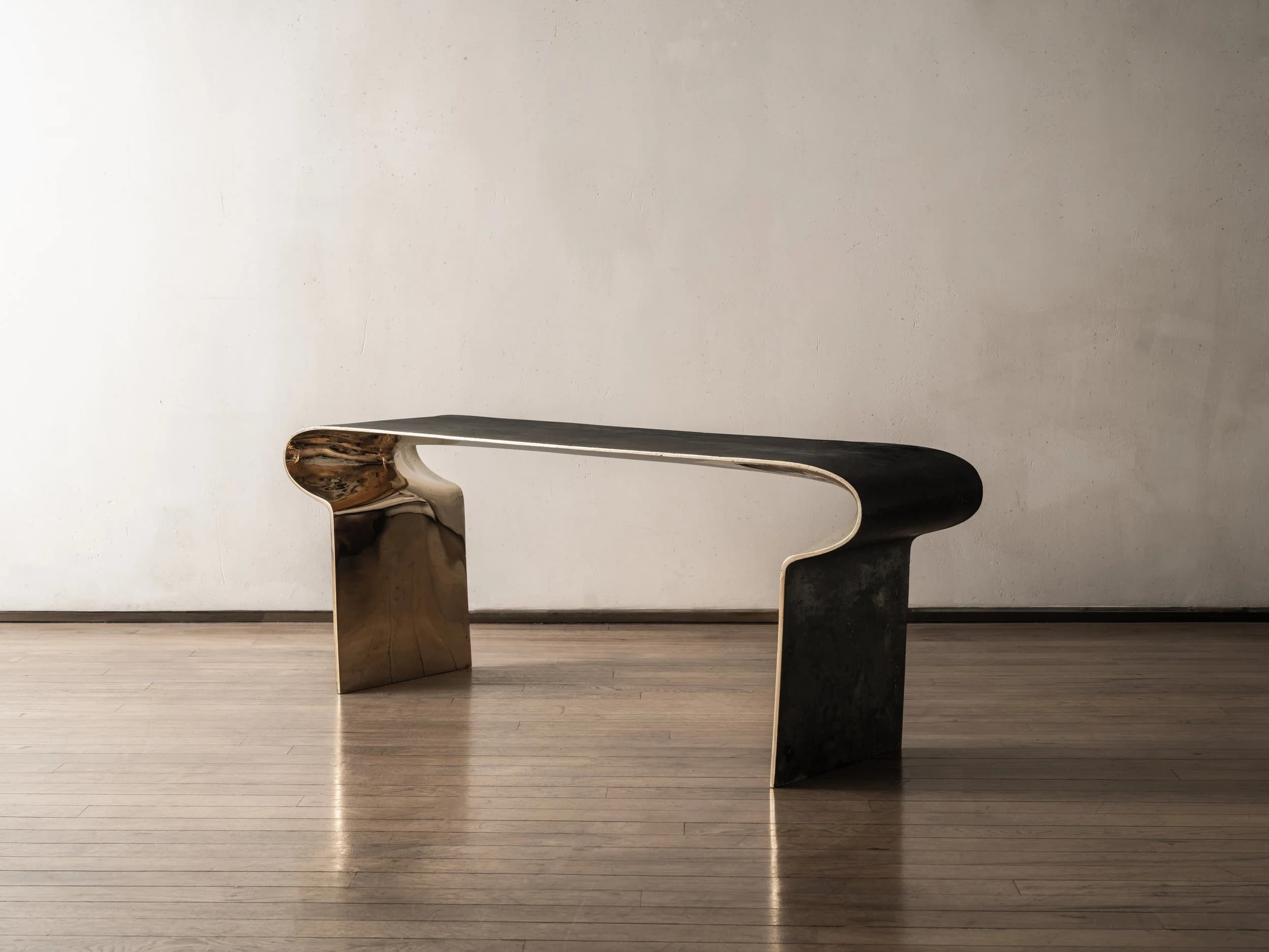 A modern sculptural console table featuring a distinctive two-tone finish. The table is formed from a single, continuous piece of material, creating a U-shaped or inverted C-shaped profile. The exterior surfaces exhibit a dark, patinated finish, while the interior curves are highly polished brass, creating a striking contrast. Its minimalist yet bold design makes it a prominent statement piece. — 3D model