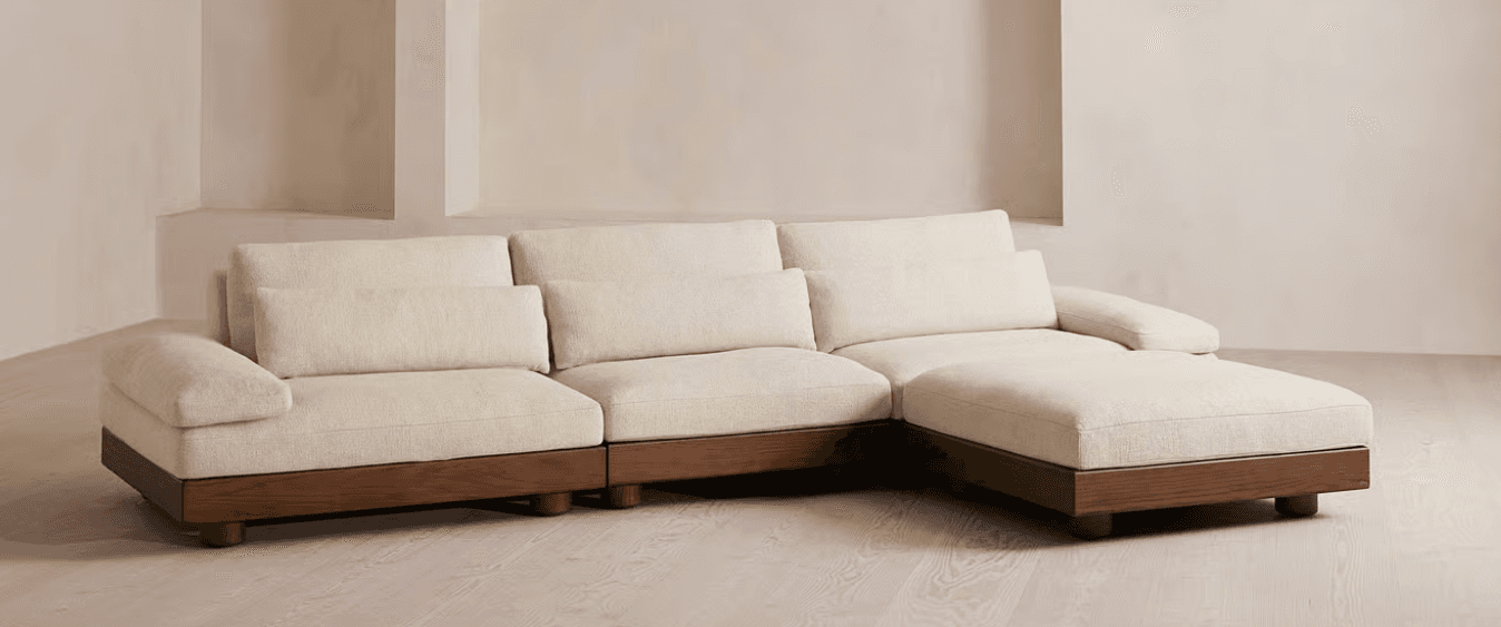 A sectional sofa featuring a modular design. It has a low profile with deep seating cushions and a chaise lounge extension on the right side. The sofa is upholstered in a textured, light-colored fabric and is supported by a dark wood base with rounded feet. — 3D model
