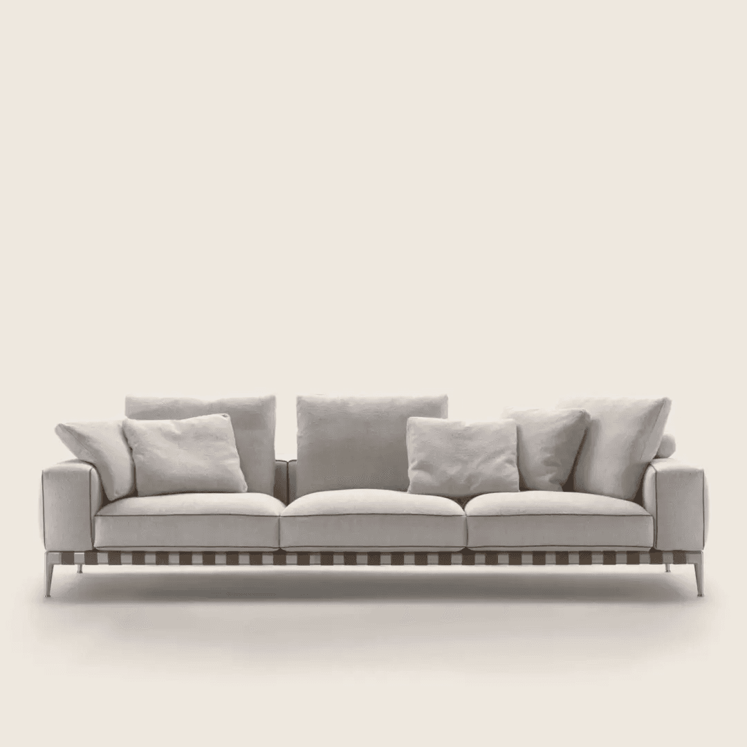 A modern three-seater sofa featuring light-colored fabric upholstery, loose back and seat cushions, and multiple throw pillows. The sofa rests on a visible wooden slatted base with a darker accent line, supported by slim, angled metal legs. It has squared arms and contrast piping along the edges of the cushions and frame. — 3D model