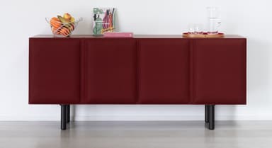 A modern, rectangular sideboard featuring a dark wood top and side panels, with three distinct front panels upholstered in a deep burgundy fabric. The upholstered panels appear to be doors, each with a subtle vertical delineation suggesting padding or channel detailing. The entire unit is supported by four slender, black cylindrical metal legs, elevating it off the floor. On its top surface rests a red circular tray holding a glass pitcher and two tumblers. — 3D model