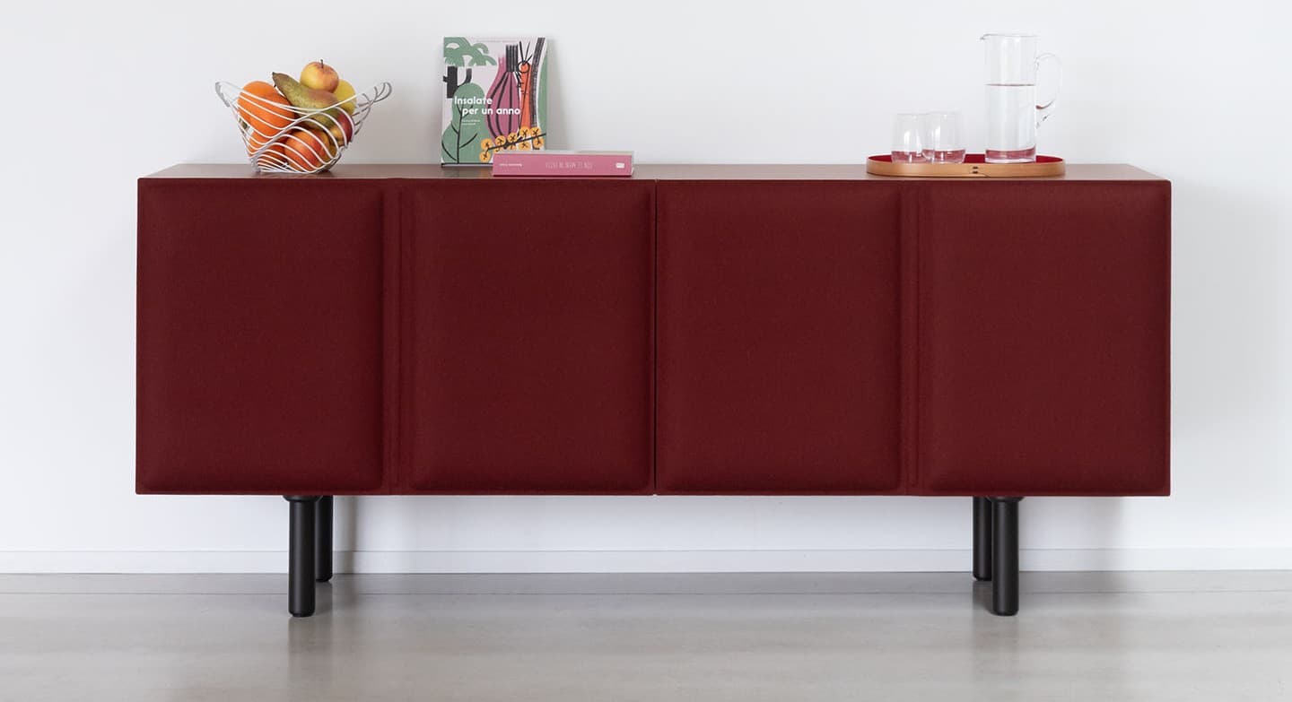 A modern, rectangular sideboard featuring a dark wood top and side panels, with three distinct front panels upholstered in a deep burgundy fabric. The upholstered panels appear to be doors, each with a subtle vertical delineation suggesting padding or channel detailing. The entire unit is supported by four slender, black cylindrical metal legs, elevating it off the floor. On its top surface rests a red circular tray holding a glass pitcher and two tumblers. — 3D model