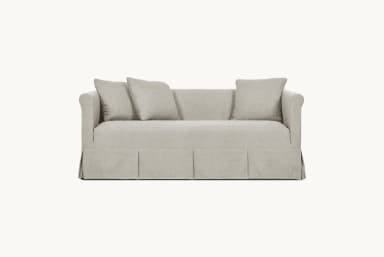 A light-colored, slipcovered sofa with a tailored skirt featuring pleats. It has plush seat and back cushions, with three throw pillows arranged on the back. The sofa arms are slightly curved. — 3D model