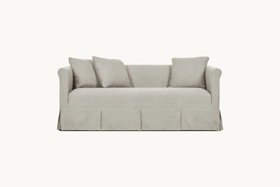A light-colored, slipcovered sofa with a tailored skirt featuring pleats. It has plush seat and back cushions, with three throw pillows arranged on the back. The sofa arms are slightly curved. — 3D model