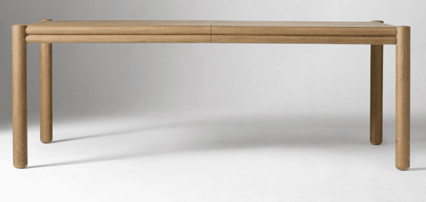 A modern, light-colored wooden dining table, likely made of oak, featuring a rectangular tabletop with a central seam suggesting extendability. It is supported by four robust cylindrical legs, one at each corner. A distinctive double-rounded apron runs horizontally beneath the tabletop, adding a sculptural detail to its minimalist design. — 3D model
