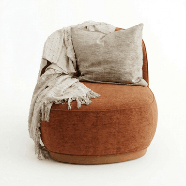 A contemporary armchair with a rounded silhouette, upholstered in a textured, rust-colored fabric. It features a low profile with a visible wooden base. A textured throw blanket with fringes and a decorative pillow are draped over the back and armrest. — 3D model