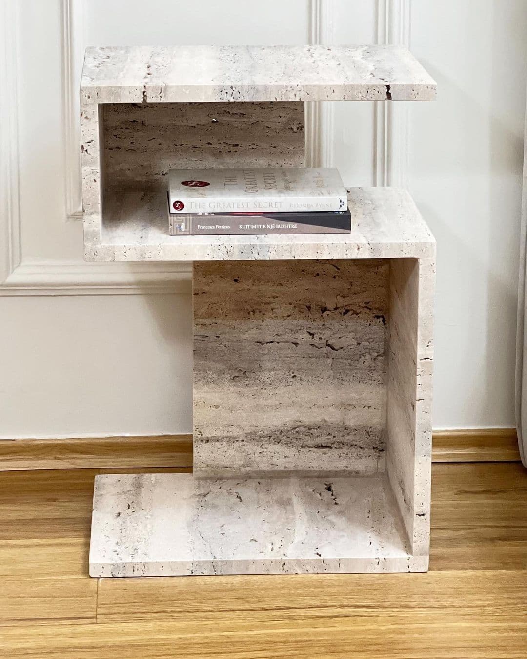A C-shaped end table constructed from travertine stone, featuring an offset stacking design. The table has two shelves, with the upper shelf cantilevered and positioned slightly forward and to the right of the lower shelf. The material exhibits natural variations in color and texture, with visible pitting and veining characteristic of travertine. Two books are placed on the upper shelf. — 3D model