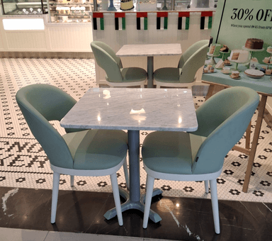 A modern dining set featuring a square table with a white marble top and a grey pedestal base, paired with two upholstered dining chairs. The chairs have light green fabric upholstery, a curved bucket-style seat and back, a small cutout in the lower back, and light-colored, slightly tapered legs. — 3D model