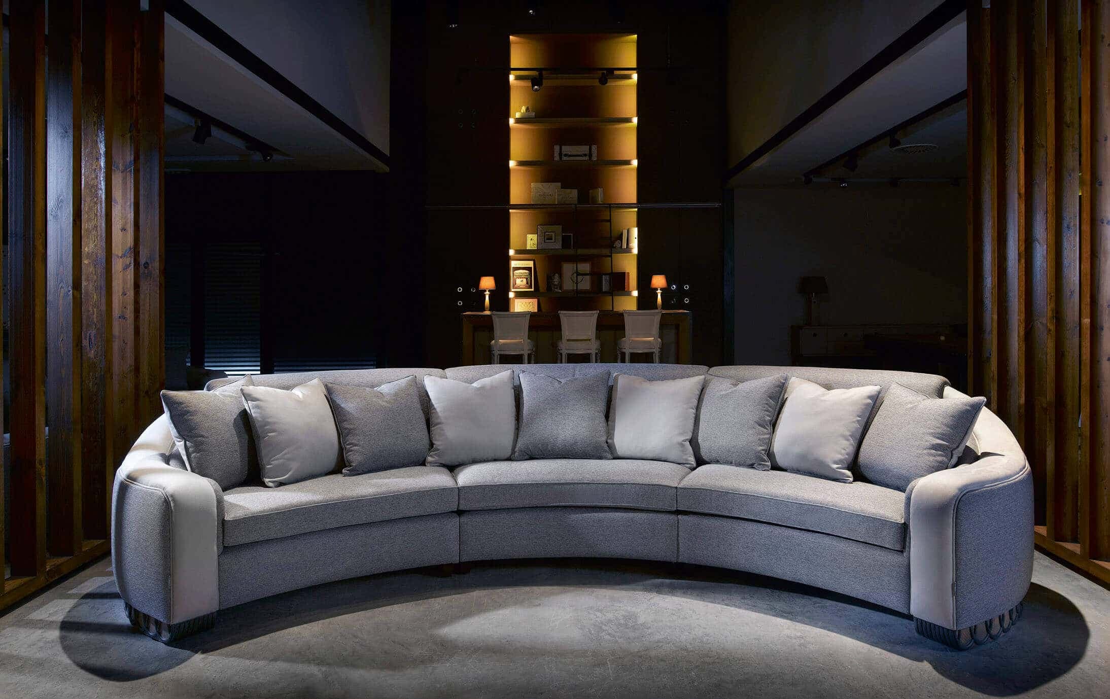 A large, contemporary curved sectional sofa upholstered in a light grey textured fabric. It features multiple loose seat and back cushions, rounded armrests with contrasting light-colored panels, and a distinctive fluted or ribbed dark base detail. — 3D model