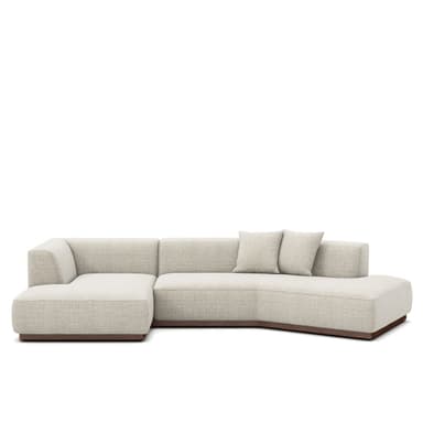 A contemporary sectional sofa featuring a light beige fabric upholstery, a low-profile design, and a dark wooden plinth base. It consists of multiple modules, including a main sofa section and an integrated chaise lounge, with subtly rounded edges. Two matching square throw pillows are included. — 3D model