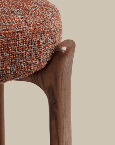 A close-up image of a modern stool, showcasing its rounded, textured upholstered seat in a reddish-brown and gray fabric, gracefully integrated with a sculpted, dark wooden leg that features a smooth, organic curve. — 3D model
