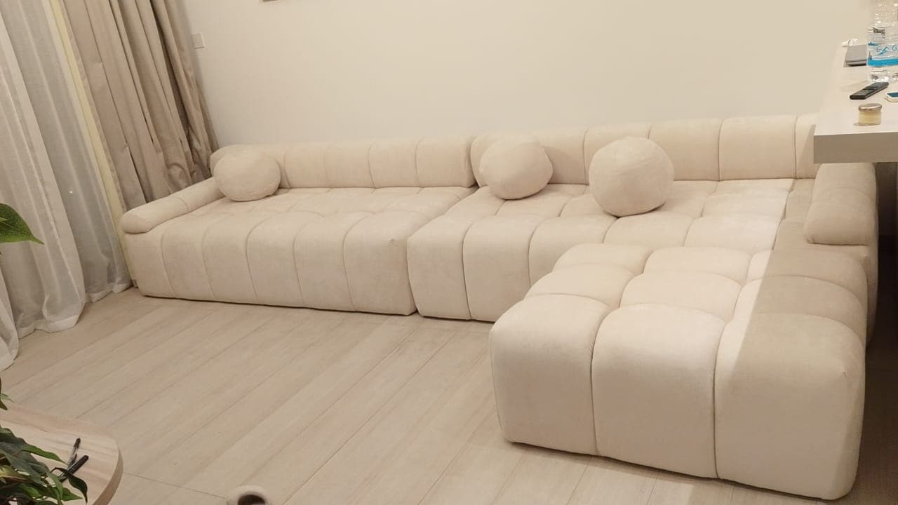A modular sectional sofa with a tufted, upholstered design. It features a light cream or off-white fabric and includes multiple square seat cushions and back cushions, along with decorative spherical throw pillows. The configuration appears to be a chaise lounge attached to a longer sofa section. — 3D model