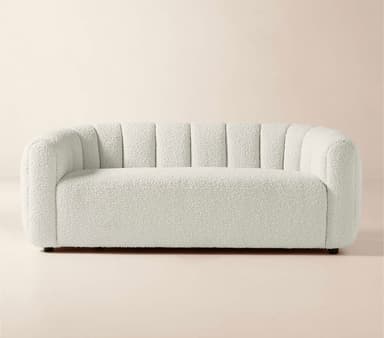 A contemporary three-seater sofa featuring a distinctive channel-tufted design on its backrest and inner arms, upholstered in an off-white bouclé fabric. The sofa has a rounded, sculptural silhouette with a low profile and recessed feet, presenting a soft and inviting texture. — 3D model