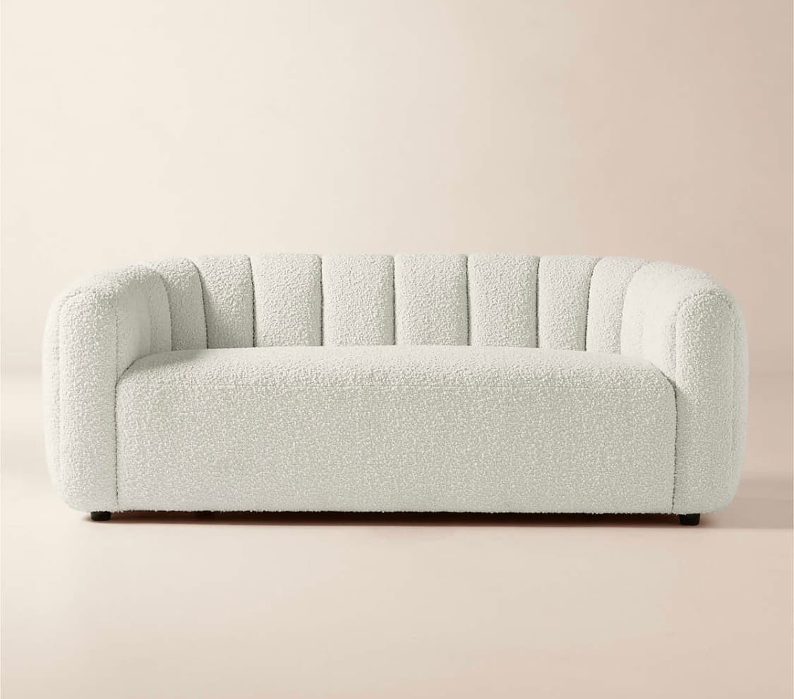 A contemporary three-seater sofa featuring a distinctive channel-tufted design on its backrest and inner arms, upholstered in an off-white bouclé fabric. The sofa has a rounded, sculptural silhouette with a low profile and recessed feet, presenting a soft and inviting texture. — 3D model
