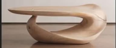 A low, sculptural coffee table characterized by its fluid, biomorphic shape crafted entirely from light-toned wood. The design features a continuous, seamless form where the tabletop gently curves downwards to create the base, eliminating distinct legs and offering a visually striking, organic silhouette. — 3D model