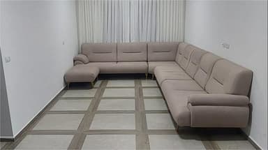 A large, U-shaped sectional sofa in a light beige fabric, featuring modular components, subtly tufted backrests, and splayed wooden legs. It is accompanied by a matching detached ottoman. — 3D model