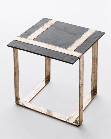 A square side table featuring a dark, textured slate top and a robust, open-cube base constructed from raw, cast bronze. The slate tabletop is inset with two perpendicular strips of the same bronze material, creating a distinctive cross pattern. The bronze frame exhibits a deliberately unfinished, organic texture, emphasizing its handcrafted nature and solid construction. The overall aesthetic is heavy, raw, and industrial, typical of Brutalist design influences. — 3D model