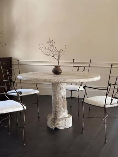 A round, pedestal table with a rough, textured surface. The pedestal has a columnar design with a decorative base and capital, reminiscent of classical architecture. A small, dark ceramic vase with a dried plant is placed on the tabletop. — 3D model