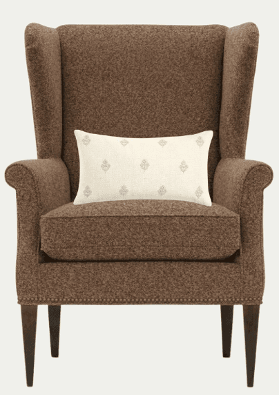 A brown tweed upholstered wingback chair with a lumbar pillow. The chair features a high back with prominent 'wings', padded armrests, and a plush seat cushion. The base is supported by four dark wooden legs, and a decorative trim of nailheads is visible along the bottom edge of the chair's frame. A rectangular lumbar pillow with a cream background and a repeating symmetrical pattern in a light brown hue is placed on the seat. — 3D model