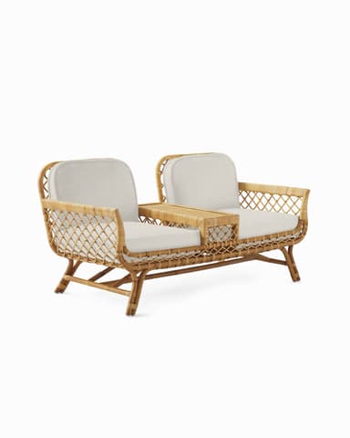 A contemporary loveseat crafted from natural rattan, featuring a woven open-patterned frame. It includes two seating areas with thick, light-colored fabric cushions, separated by a rectangular integrated table section made of solid rattan. The frame showcases curved lines and sturdy legs, presenting a light and airy aesthetic suitable for various relaxed settings. — 3D model