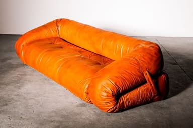 A vintage or vintage-style sofa featuring a plush, rounded, and deeply tufted design, upholstered in a rich cognac-colored leather. The sofa has a distinctive 'puffy' appearance, with a backrest and seat formed from large, soft, cylindrical sections. Straps with button details are visible on the side, suggesting a modular construction or adjustable elements. The overall aesthetic is comfortable and relaxed, reminiscent of iconic Italian designs from the 1970s. — 3D model