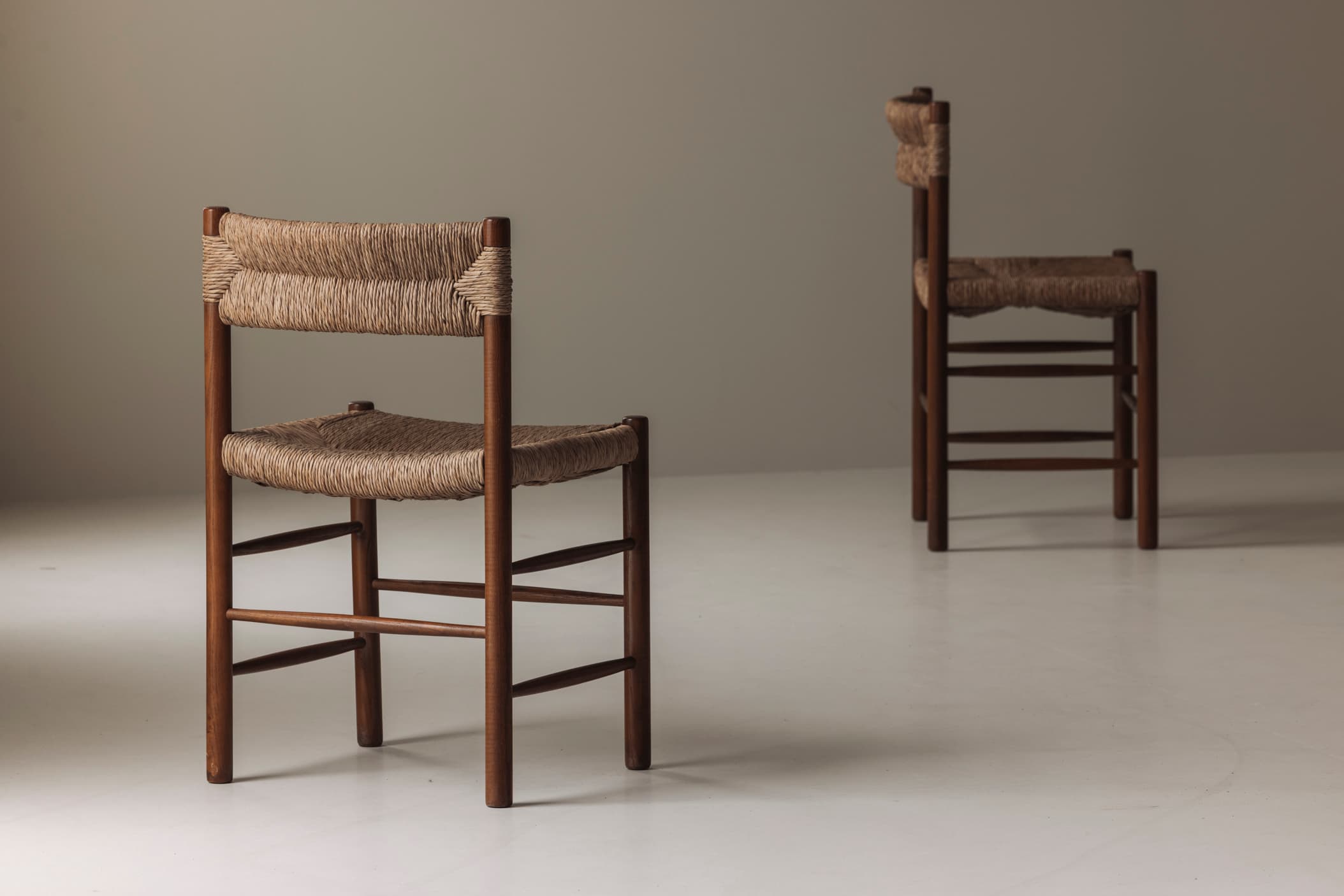 A pair of armless chairs featuring a sturdy wooden frame and intricately woven cord for both the seat and backrest. The chairs display a simple, functional design with round dowel stretchers connecting the legs. The wood appears to be a medium to dark brown stain, complementing the natural beige tone of the woven material. The overall aesthetic is one of durability and organic texture. — 3D model