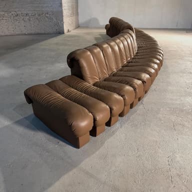 A modular, curved sectional sofa upholstered in brown leather. The sofa features a distinctive design with vertical channel tufting on the seat and back cushions, creating a segmented appearance. It is composed of multiple individual units that form a large, L-shaped seating arrangement with an additional projecting section. — 3D model