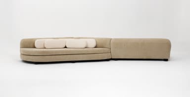 A contemporary sinuous modular sofa featuring a serpentine form, upholstered in a beige textured fabric. The design includes integrated cylindrical backrests/armrests and is accompanied by smaller, contrasting cream-colored cylindrical lumbar pillows. The sofa appears to be composed of two interconnected, curved modules. — 3D model