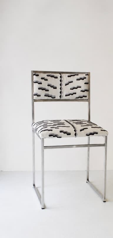 A contemporary armless chair featuring a minimalist, rectangular frame constructed from polished tubular metal. The seat and backrest are upholstered in a fabric with an abstract geometric pattern in shades of cream, black, and grey. The frame forms continuous loops for the legs and supports the seat and back. — 3D model
