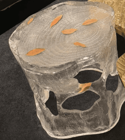 A sculptural accent table or stool crafted from clear, textured resin, designed to resemble a block of ice or a natural tree stump. The piece features an organic, irregular silhouette with cut-out sections in its body, giving it a light, ethereal appearance. Embedded within the clear material are small, delicate golden leaf inclusions. The top surface is flat but textured with an embossed wood grain pattern, suggesting tree rings, further enhancing its nature-inspired aesthetic. The overall impression is one of modern artistry combined with natural elements. — 3D model