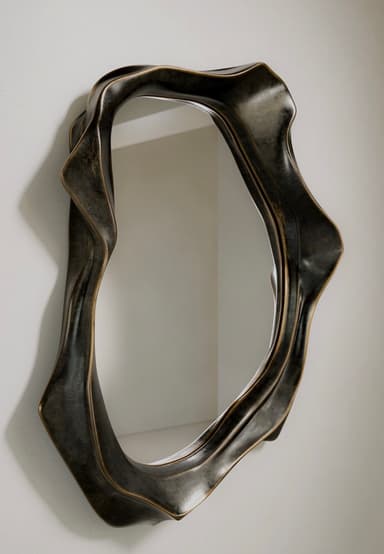 A wall-mounted mirror featuring an irregularly shaped, organic, and sculptural frame. The frame has a dark, aged metallic finish, possibly bronze or antique brass, with textured, undulating surfaces that create a sense of movement and depth. The mirror glass itself is also irregularly shaped, conforming to the inner edge of the fluid frame. It casts a soft shadow on the white background, emphasizing its three-dimensional form. — 3D model