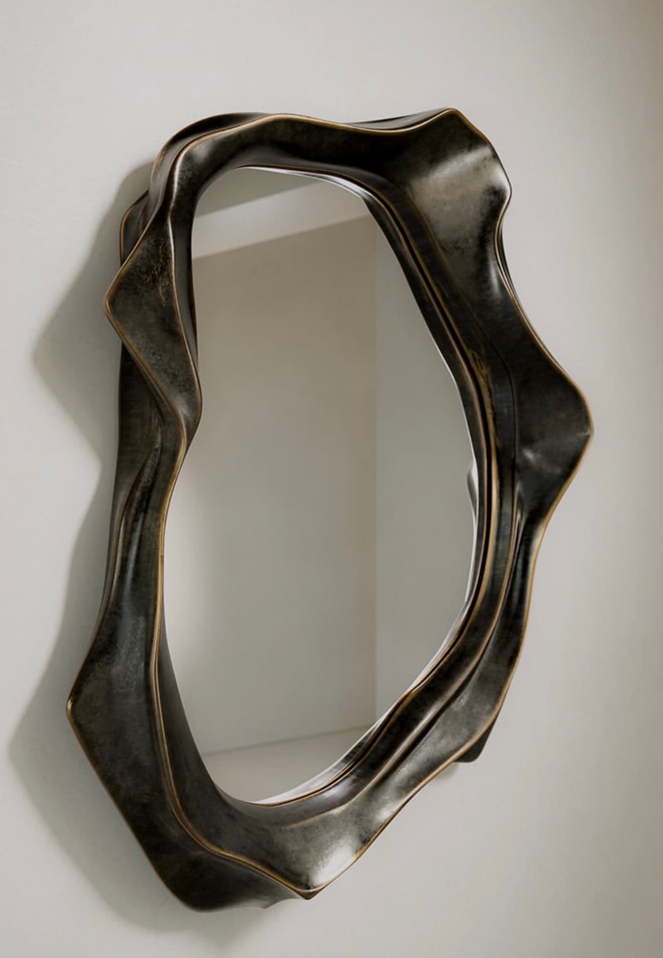 A wall-mounted mirror featuring an irregularly shaped, organic, and sculptural frame. The frame has a dark, aged metallic finish, possibly bronze or antique brass, with textured, undulating surfaces that create a sense of movement and depth. The mirror glass itself is also irregularly shaped, conforming to the inner edge of the fluid frame. It casts a soft shadow on the white background, emphasizing its three-dimensional form. — 3D model