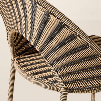 A close-up image of a contemporary accent chair constructed from two-tone synthetic rattan. The chair features a tightly woven pattern in alternating bands of black and light beige, creating a striped effect. It has a curved backrest and integrated armrests, suggesting a comfortable, ergonomic form. The legs, visible at the bottom, also appear to be wrapped in the same woven material, indicating a cohesive design. The texture is smooth and uniform, characteristic of synthetic materials. — 3D model