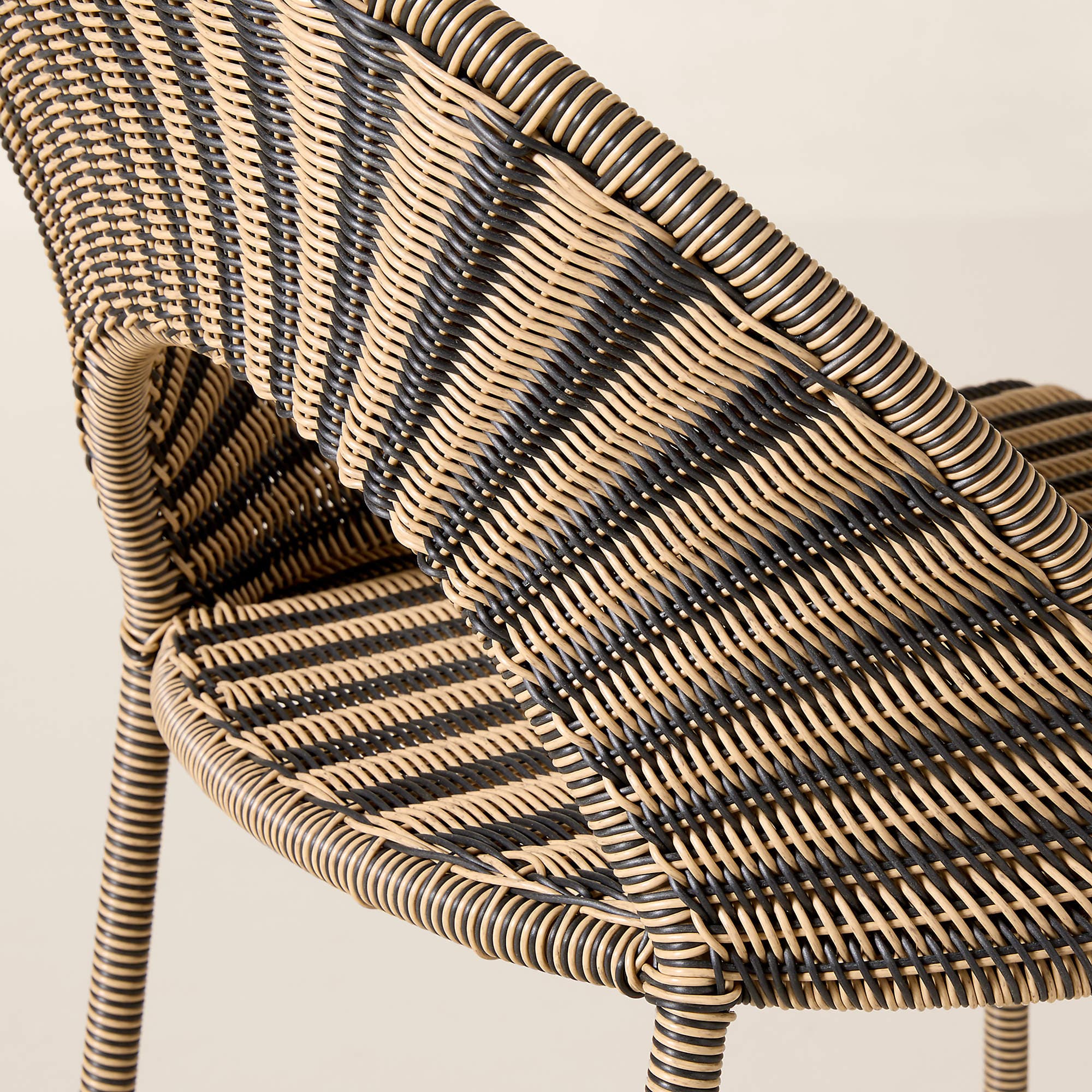 A close-up image of a contemporary accent chair constructed from two-tone synthetic rattan. The chair features a tightly woven pattern in alternating bands of black and light beige, creating a striped effect. It has a curved backrest and integrated armrests, suggesting a comfortable, ergonomic form. The legs, visible at the bottom, also appear to be wrapped in the same woven material, indicating a cohesive design. The texture is smooth and uniform, characteristic of synthetic materials. — 3D model