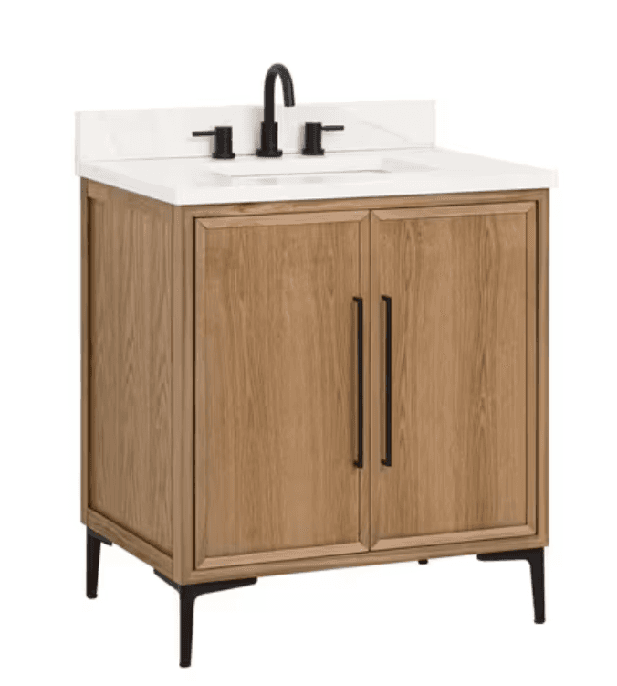 A modern freestanding bathroom vanity featuring a light natural oak-look cabinet with two recessed panel doors. It is topped with a white faux marble or quartz countertop, which includes an integrated rectangular sink and a pre-installed matte black widespread faucet. The vanity stands on four sleek, tapered matte black metal legs, complementing the door handles and faucet. — 3D model
