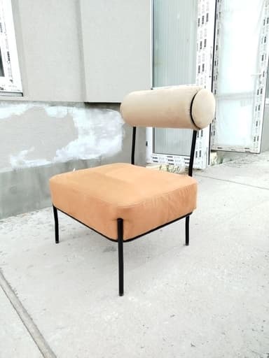 A modern armless lounge chair with a low-profile, rectangular cushioned seat in light brown/tan faux leather, supported by a minimalist black metal frame. It features a distinctive cylindrical backrest upholstered in light beige fabric, attached to the frame. — 3D model