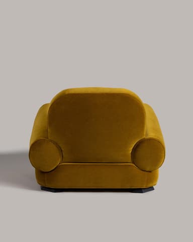 A compact, rounded armchair upholstered in a mustard-yellow velvet fabric. The armchair features a high, curved backrest that seamlessly transitions into padded armrests. The seat is low and broad, with a plain upholstered base. The overall design is plush and inviting, with a visible seam line indicating the upholstery construction. — 3D model
