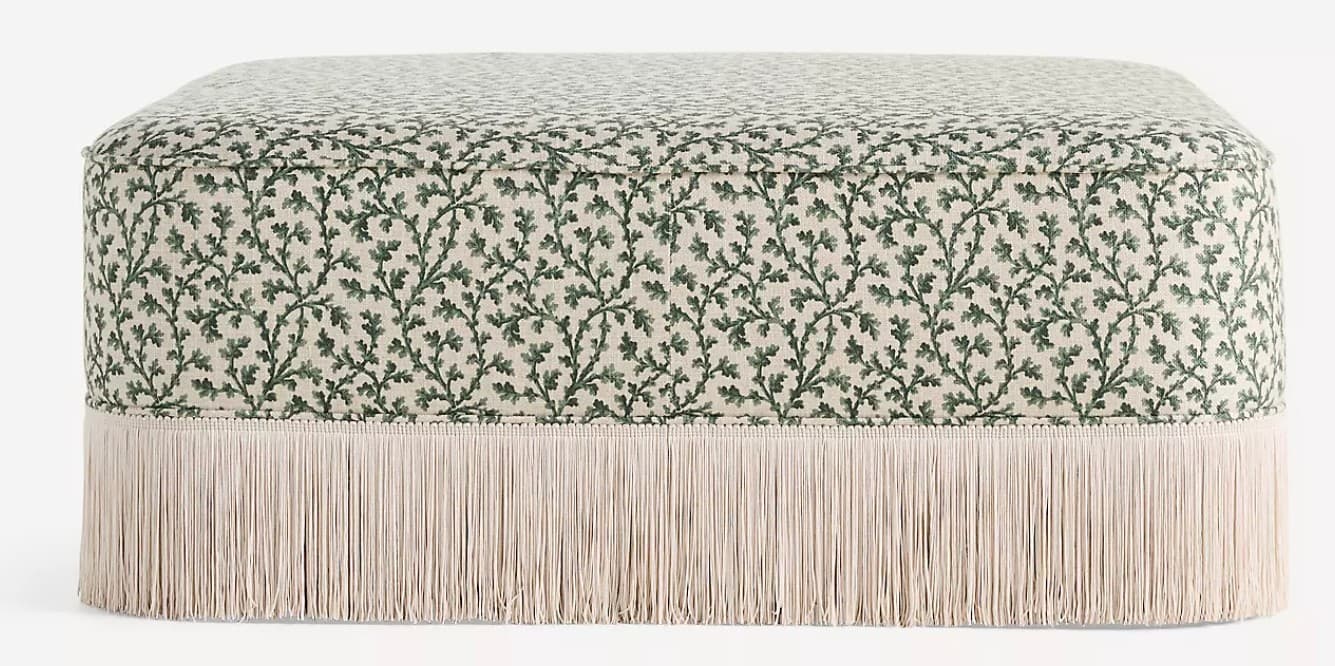 A rectangular ottoman with a padded top, upholstered in a fabric featuring a repeating pattern of green oak leaves and branches on a cream background. The base of the ottoman is adorned with a thick fringe of cream-colored tassels. — 3D model